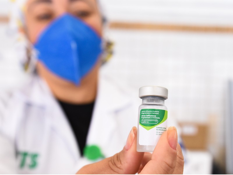 Foto de profissional da saúde segurando um frasco da vacina contra influenza. Em primeiro plano aparece a mão da profissional segurando o frasco, que possui rótulo branco e verde. A pessoa ao fundo está com máscara azul, jaleco branco e blusa preta por baixo e está desfocada